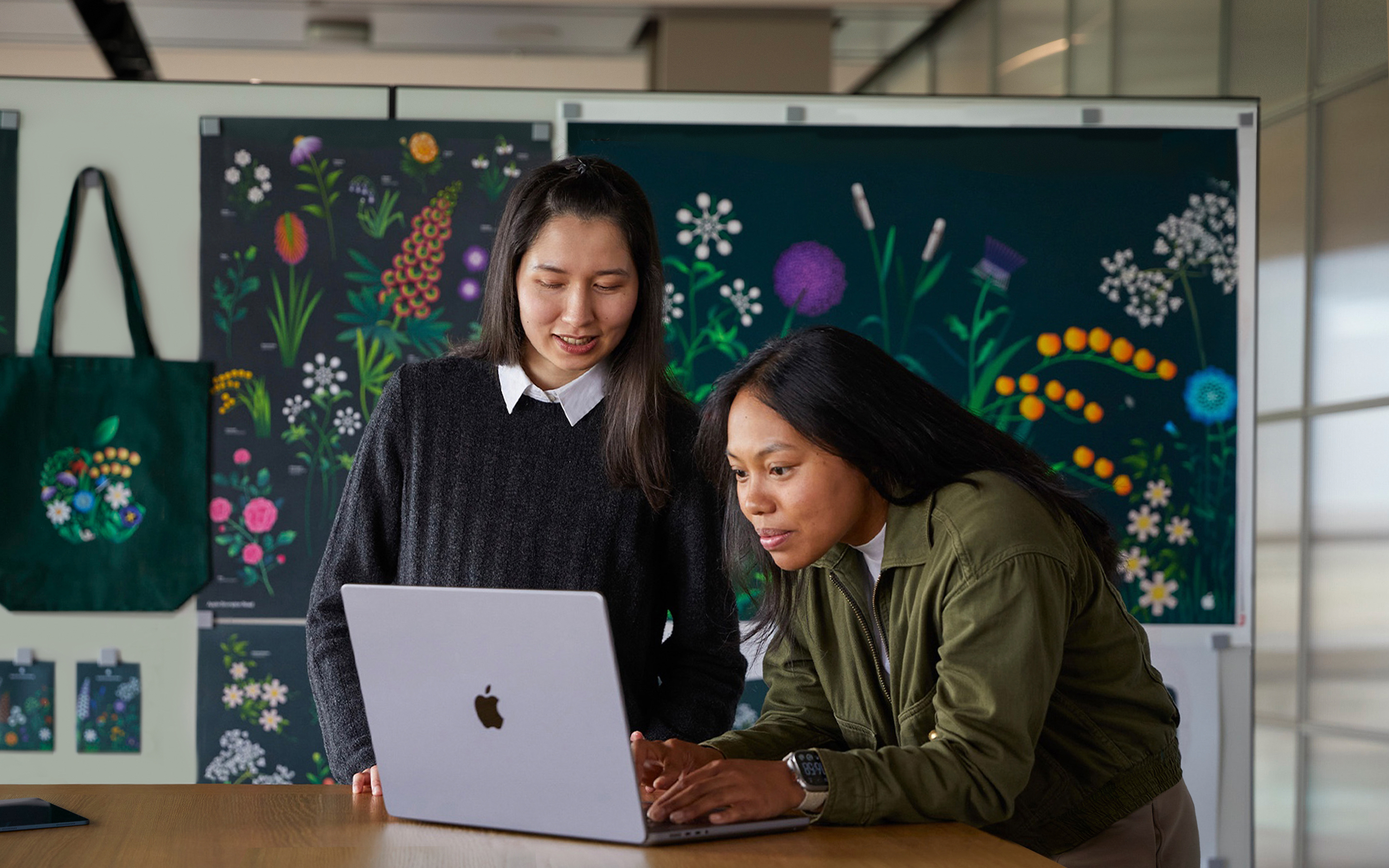 Zwei Mitglieder des Apple Marketing Teams arbeiten gemeinsam an einem MacBook vor Postern, die ihr Team designt hat.