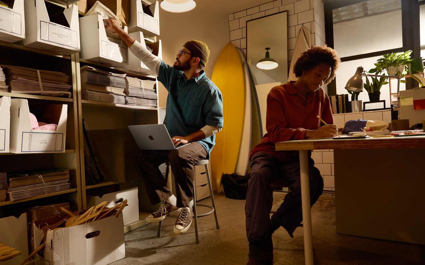 Una persona sentada en un escritorio usa un lápiz para escribir en un cuaderno, otra persona sentada junto a un estante tiene un MacBook en el regazo y estira el brazo derecho para alcanzar un archivo.