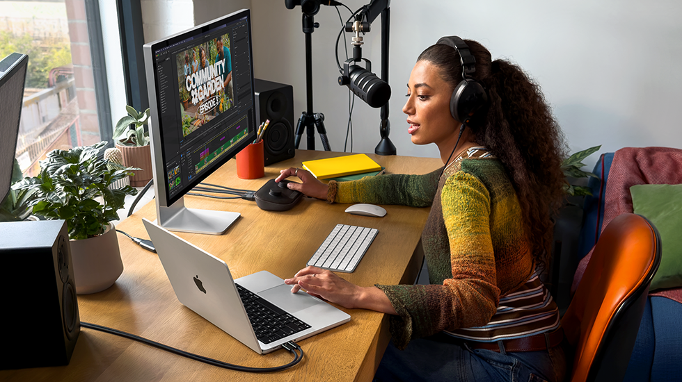 Žena sedí doma u pracovního stolu a používá MacBook Pro připojený k externímu monitoru, má nasazená sluchátka a mluví do mikrofonu
