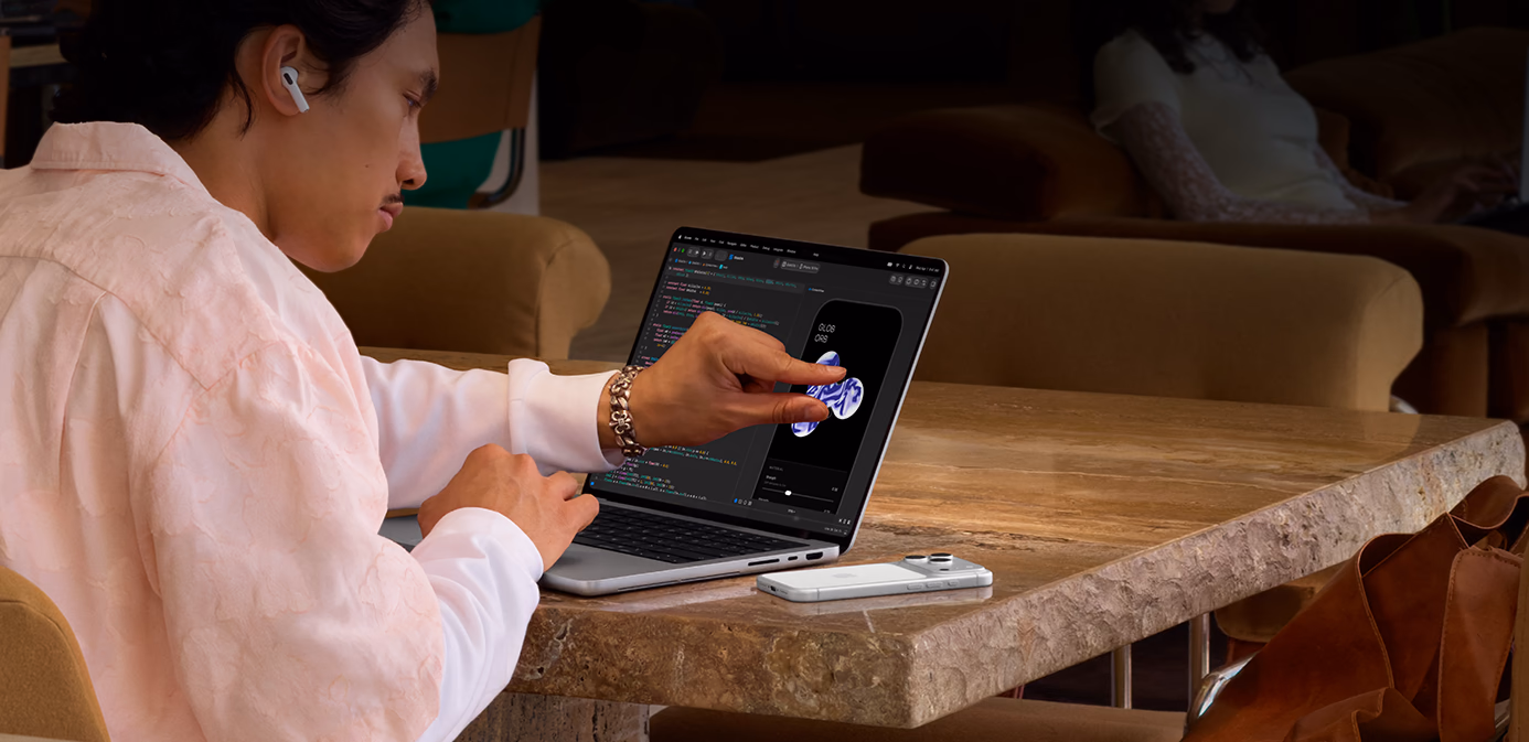 Seseorang sedang duduk di dekat meja di ruang publik, menggunakan gerakan mencubit untuk menyesuaikan elemen di Xcode dengan fitur Sentuh pada MacBook Pro berwarna perak dengan Layar PROLED XDR, sebuah iPhone terletak di atas meja di samping laptop, dan orang tersebut sedang menggunakan AirPods Pro