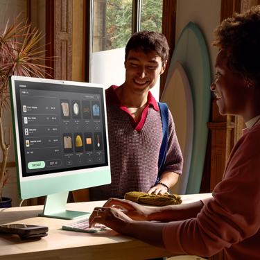 Una persona, sentada detrás del mostrador de una tienda que vende tablas de surf y otros artículos, usa una iMac para completar la compra de un cliente que está del otro lado del mostrador.