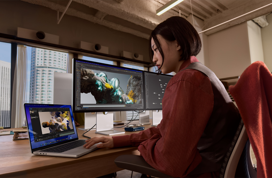 A person works at a desk with a MacBook Pro connected to external display, both screens showing a 3D render, buildings visible through the window behind