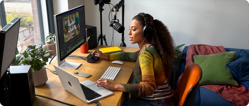 امرأة في منزلها تعمل خلف مكتبها على MacBook Pro متصل بشاشتين خارجيتين وترتدي سماعات رأس وتتحدث في الميكروفون