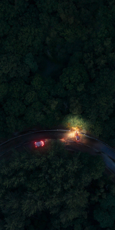 An overhead view of a winding road at night, two vehicles perpendicular with headlines on, surrounded by trees