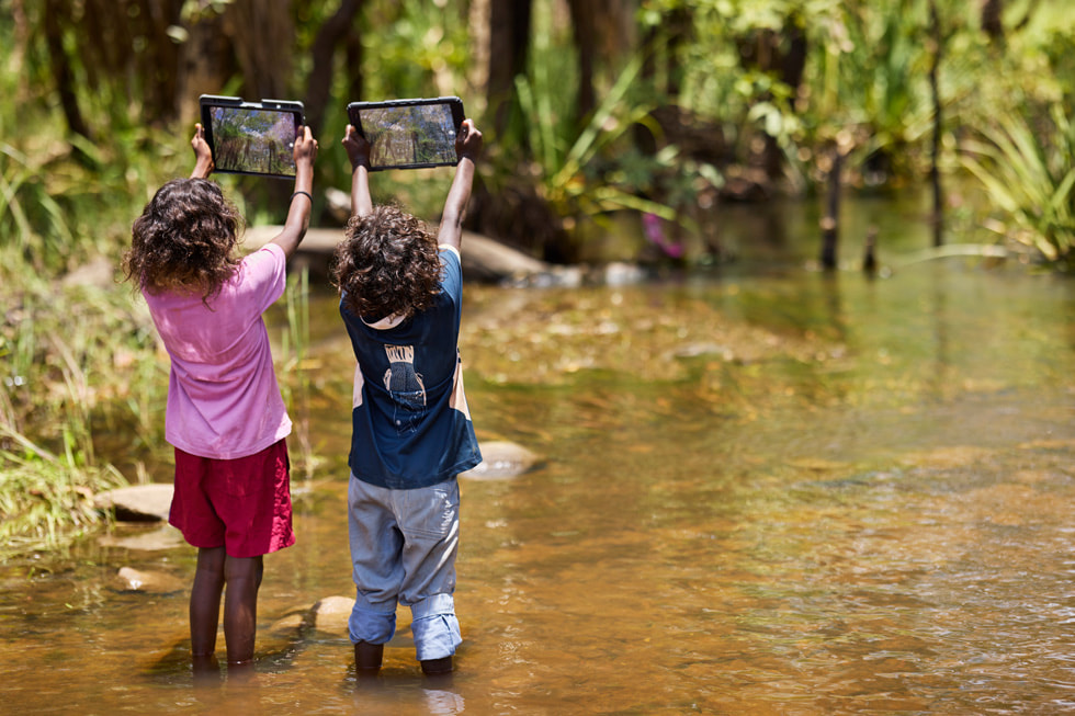 Two students from Mamadawerre in Arnhem Land hold up two iPad devices outdoors while wading in shallow water.