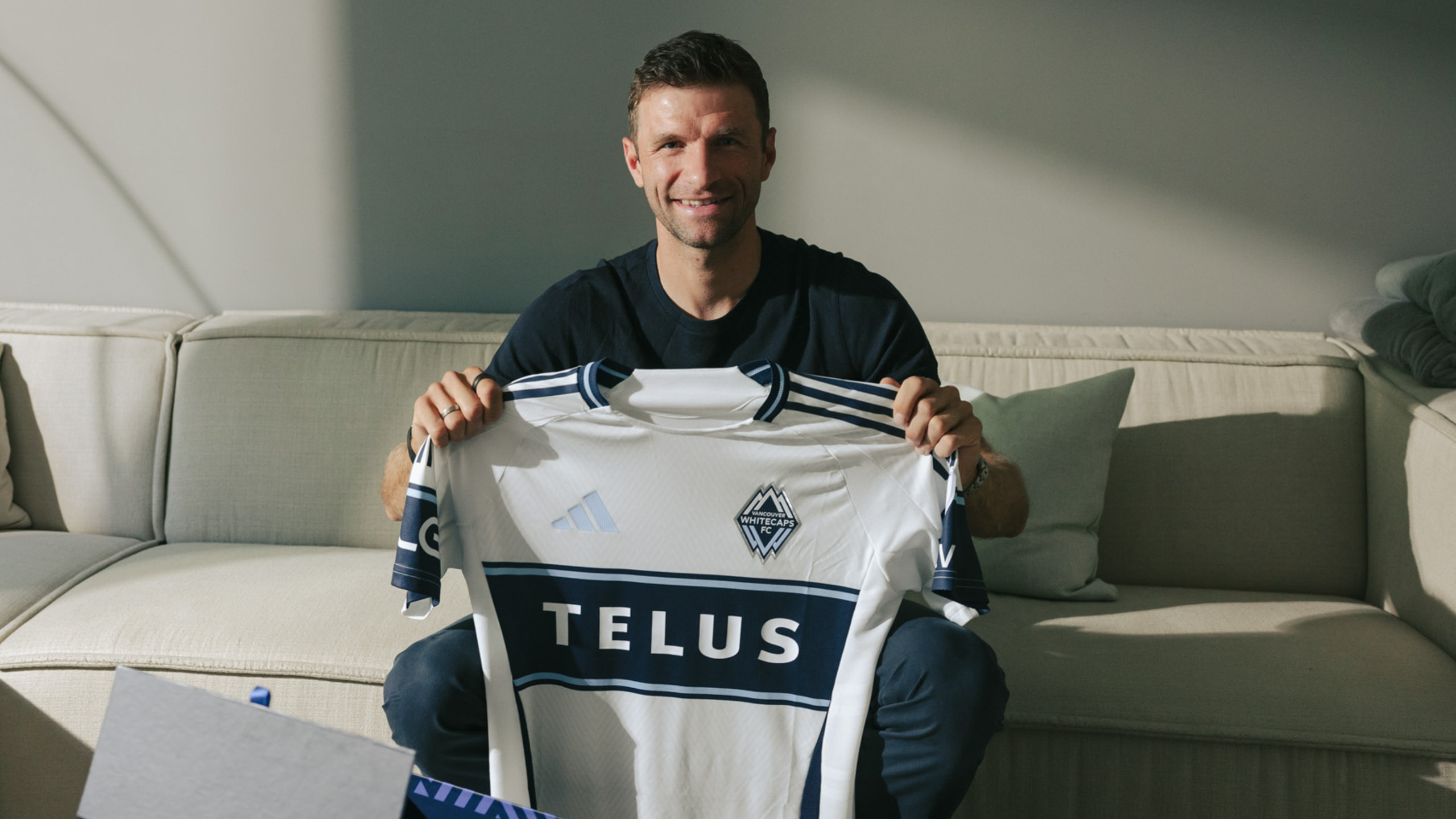 Thomas Müller sitzt auf einer Couch und zeigt ein neues Trikot des Vancouver Whitecaps FC.
