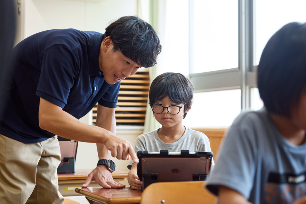 京都市の小学校の教室で、机でiPadを使って子どもと一緒に作業する教師。
