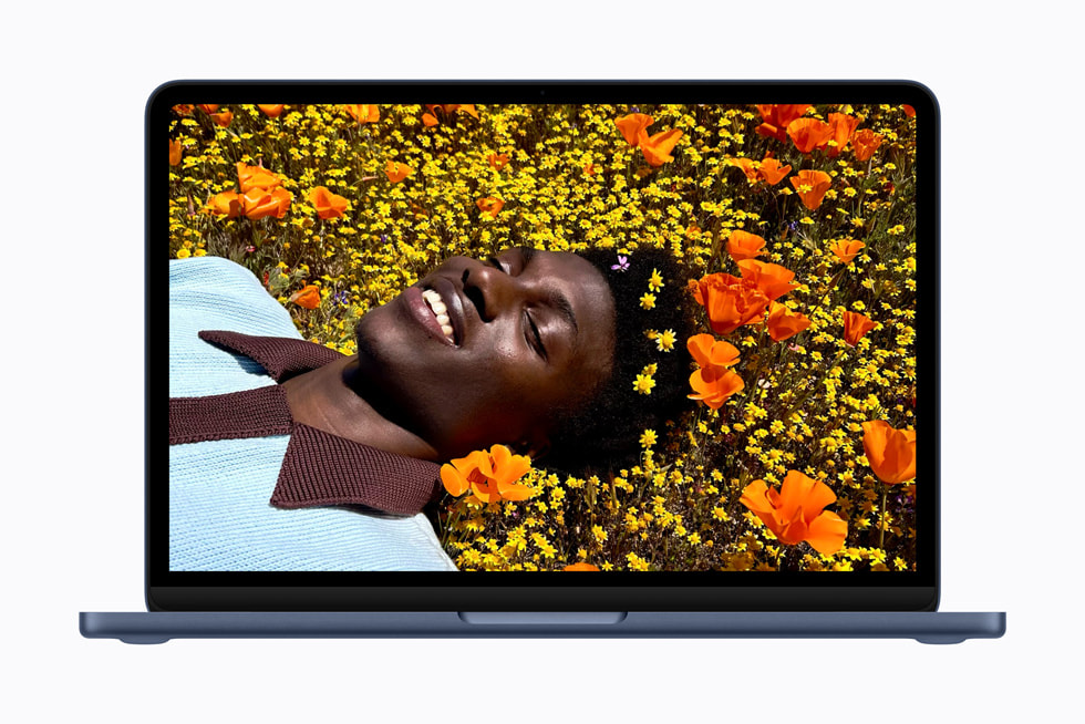 An open MacBook Neo in indigo shows a vibrant photo of a person laying in a field of flowers.