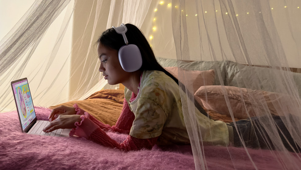 A person lounges in bed using MacBook Neo while listening to music on AirPods Max.