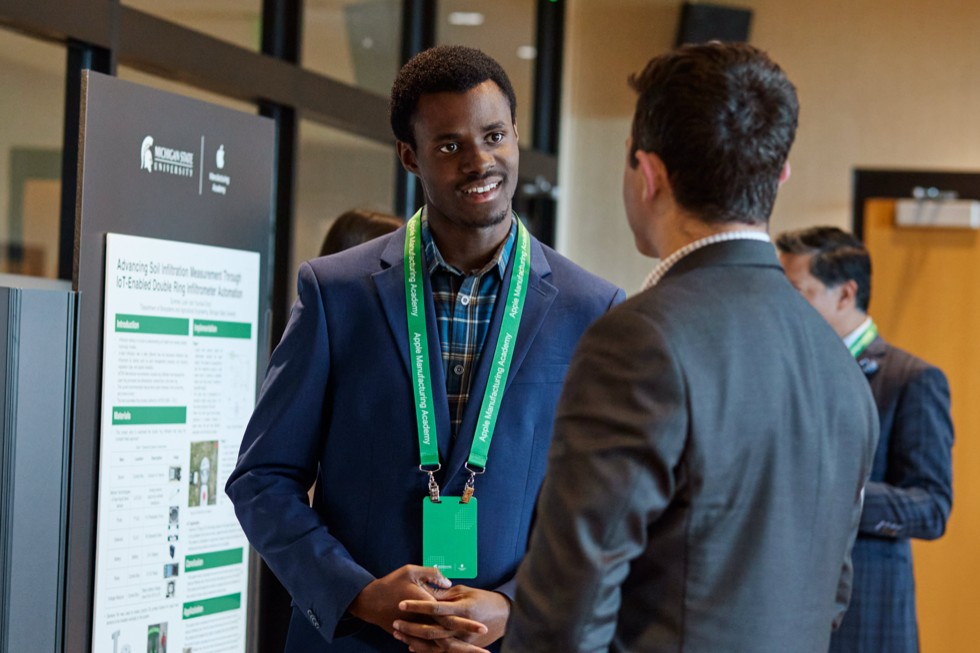 Two attendees speak while standing in front of a poster board at the Apple Manufacturing Academy’s Spring Forum.
