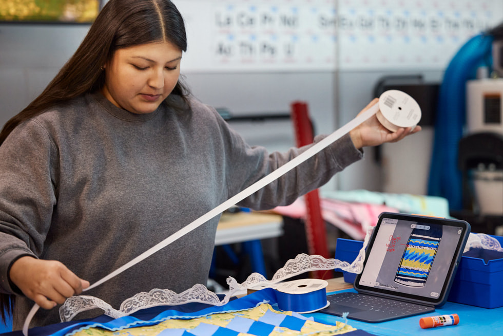 A student is shown holding ribbon while working at a station with a propped-up iPad that features a design on the screen.