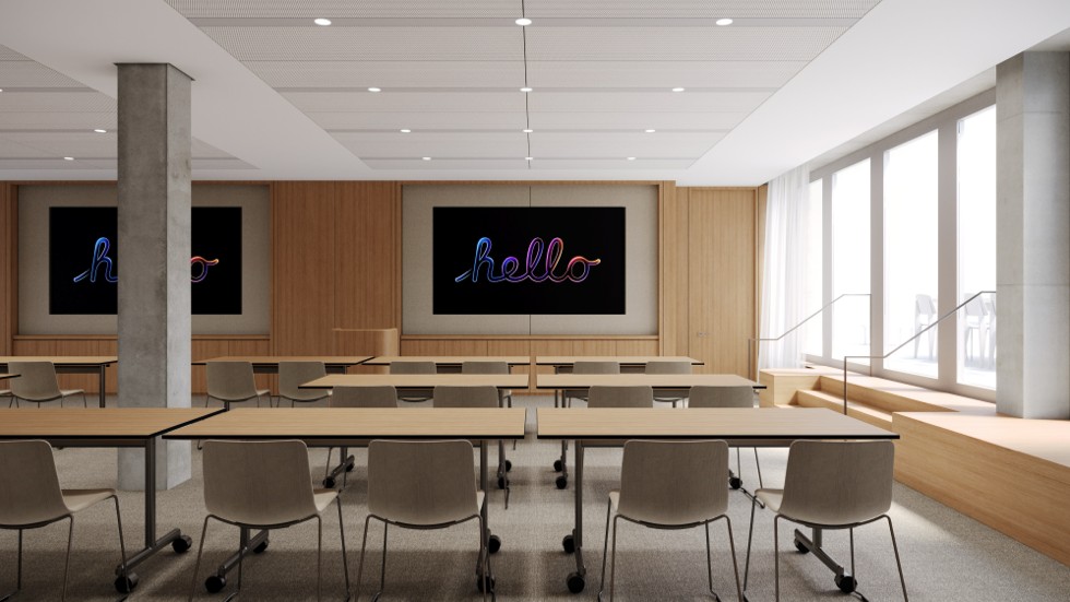 Ein leerer Schulungsraum ist mit Tischen und Stühlen ausgestattet, die auf zwei grosse Bildschirme an einer holzvertäfelten Wand ausgerichtet sind, auf denen jeweils das Wort „hello“ in kursiver Neonschrift angezeigt wird.
