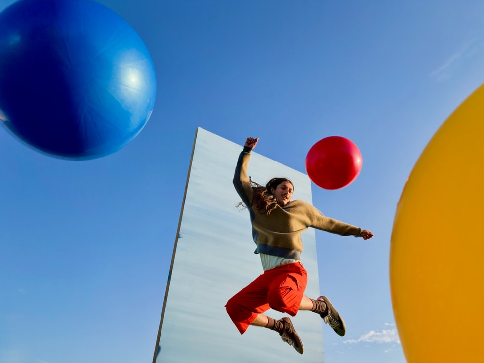 Outdoors, a person jumps as colorful inflated balls are tossed in the air against a blue sky and a rectangular gray backdrop.