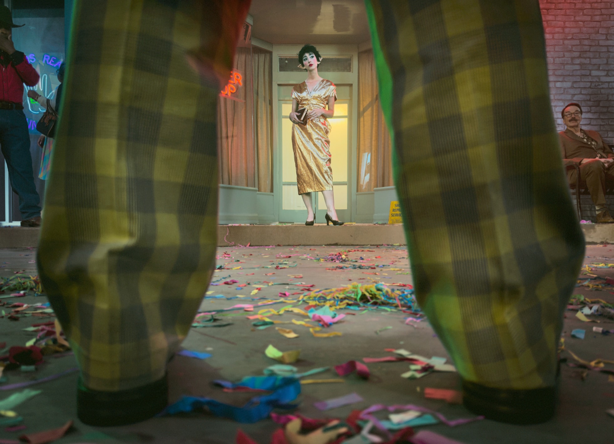 In the foreground, two legs wearing plaid pants are shown among a confetti-strewn ground, while a second subject in a gold dress stands in the background.