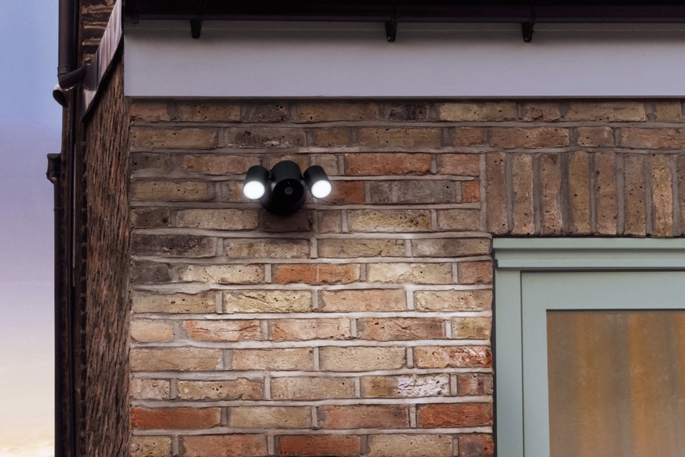 Floodlight Cam in Essence Graphite is mounted on a brick wall above a door, illuminating the area at night.