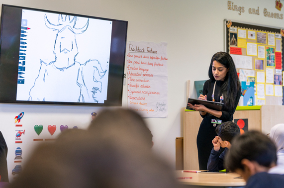 Teacher with iPad in front of classroom.