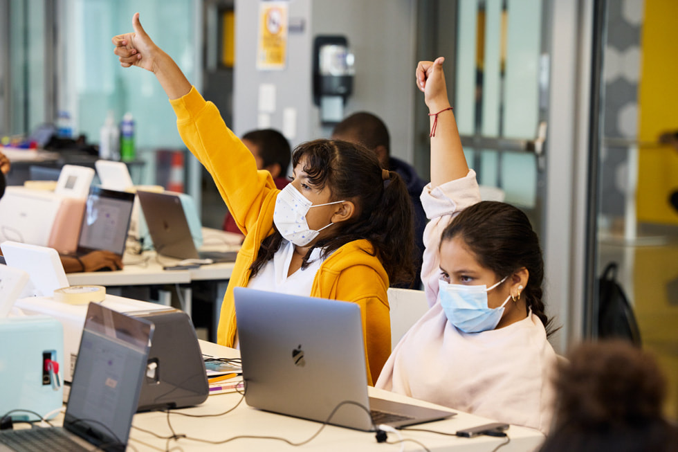 Two students using Apple notebooks raise their hands in a classroom environment.