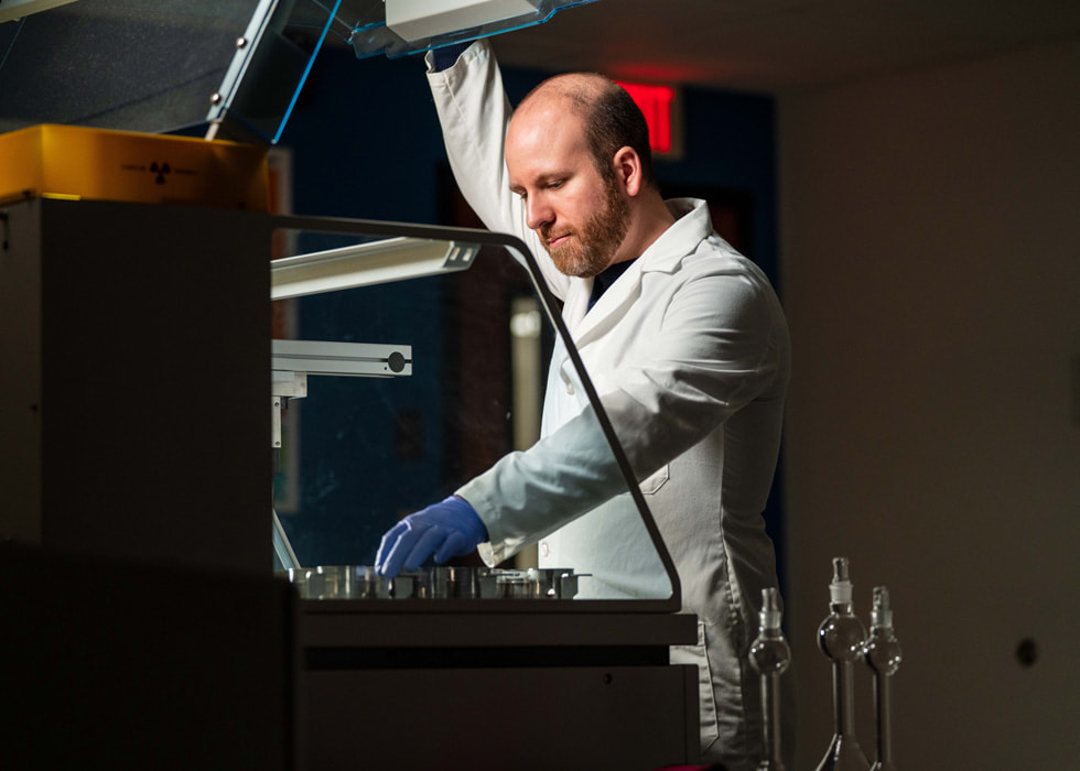 A man in a lab coat and gloves works with machinery in a lab setting.