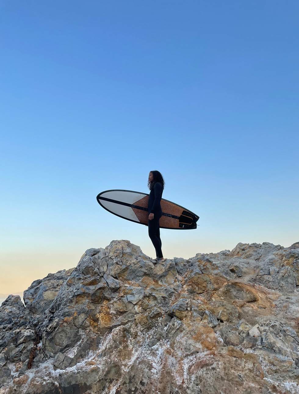 Marikah with surfboard, standing on a rock ledge.