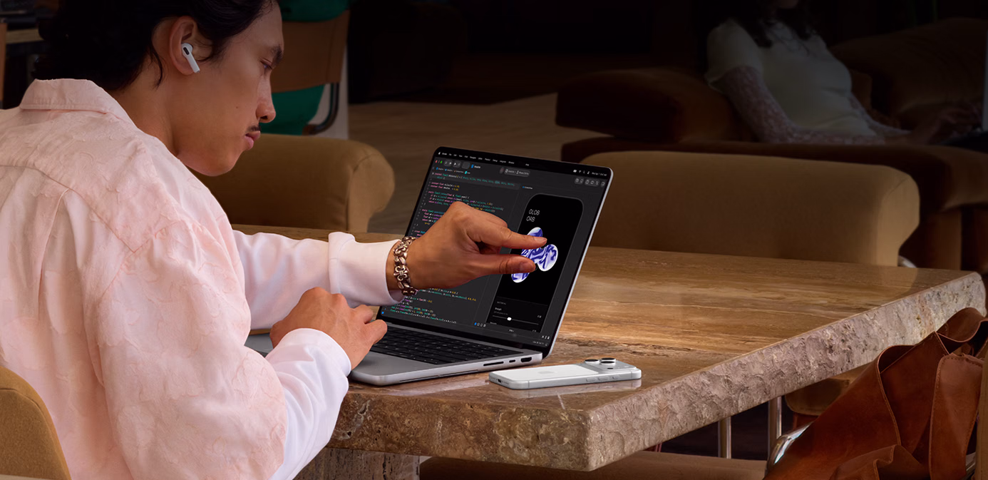 Osoba siedzi przy stole w przestrzeni publicznej i gestem szczypnięcia dostosowuje element w Xcode, korzystając z funkcji Dotyk na srebrnym MacBooku Pro z wyświetlaczem PROLED XDR; obok laptopa na stole leży iPhone, a osoba ma w uszach słuchawki AirPods Pro