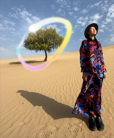 Picture of a woman in a multicoloured dress standing in a desert landscape with a lone tree in the background circled in a glowing rainbow gradient showcasing Apple Intelligence capability to erase background objects