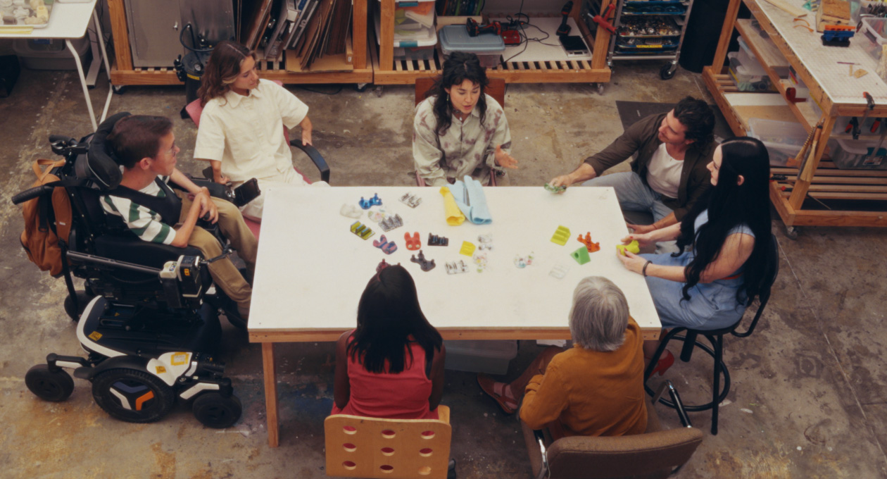 Un groupe de personnes ayant divers besoins en matière de mobilité qui se réunissent autour d’une table pour échanger sur une variété d’accessoires colorés pour smartphones, conçus pour répondre à leurs besoins spécifiques