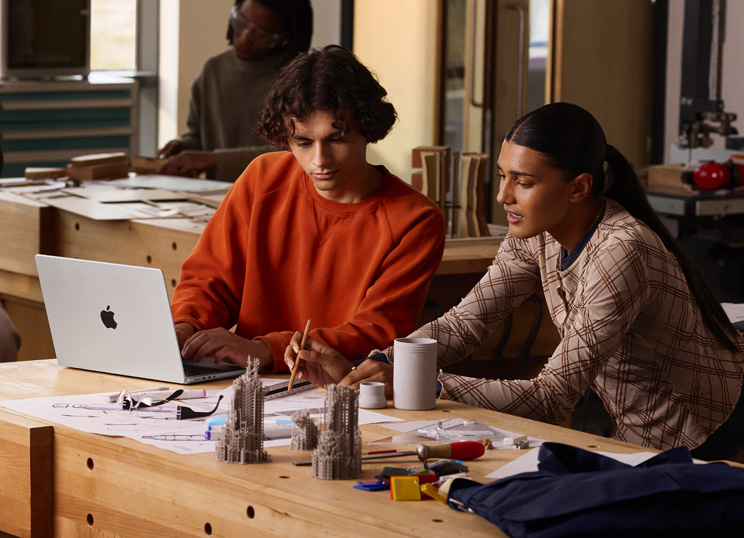Due persone che collaborano a un progetto a un tavolo, una sta digitando su un MacBook, l’altra sta usando una matita e un righello su carta, circondata da materiale artistico.