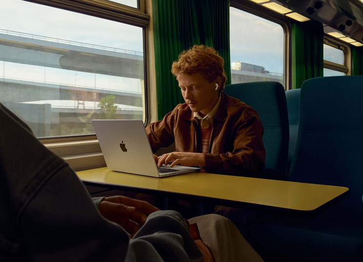 Una persona in treno seduta accanto al finestrino che usa degli AirPods e lavora su un MacBook.