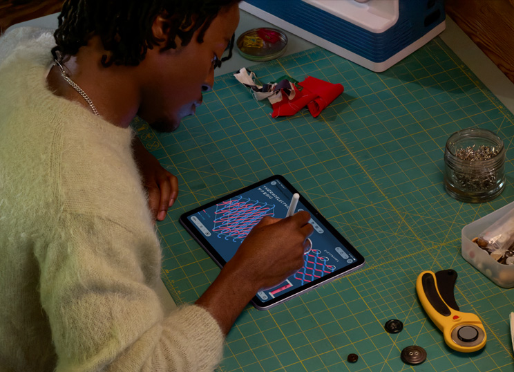 Una persona che fa un disegno con Apple Pencil su iPad, seduta davanti a un tappetino da taglio verde con materiale da cucito.