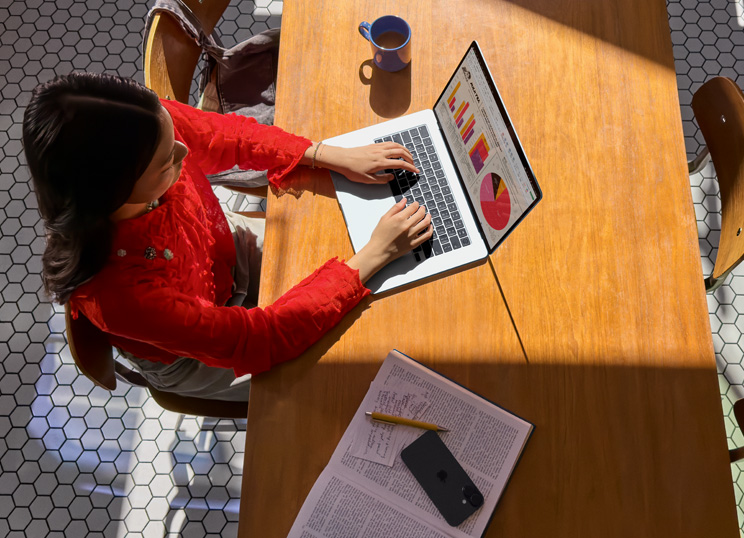 Una persona che usa un MacBook a un tavolo, con una tazza, uno smartphone e dei documenti intorno.