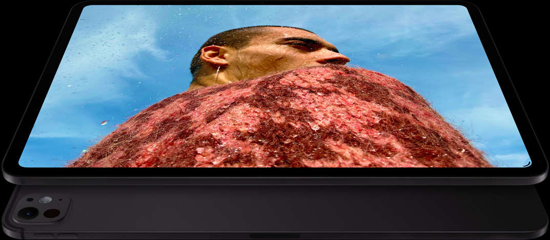 Un iPad Pro, parte frontal en ángulo, posición horizontal, color negro espacial, esquinas redondeadas, marco de la pantalla negro, la pantalla muestra la foto de un hombre con un suéter rojo bajo un cielo azul, debajo, otro iPad Pro, parte posterior, posición horizontal, color negro espacial, sistema de cámaras Pro, botón de subir volumen, botón de bajar volumen
