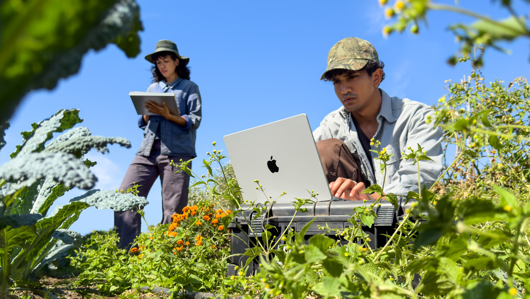 Dos personas trabajando en un jardín, una usa una MacBook Pro color plata que no está conectada a la corriente y la otra sostiene un bloc de notas