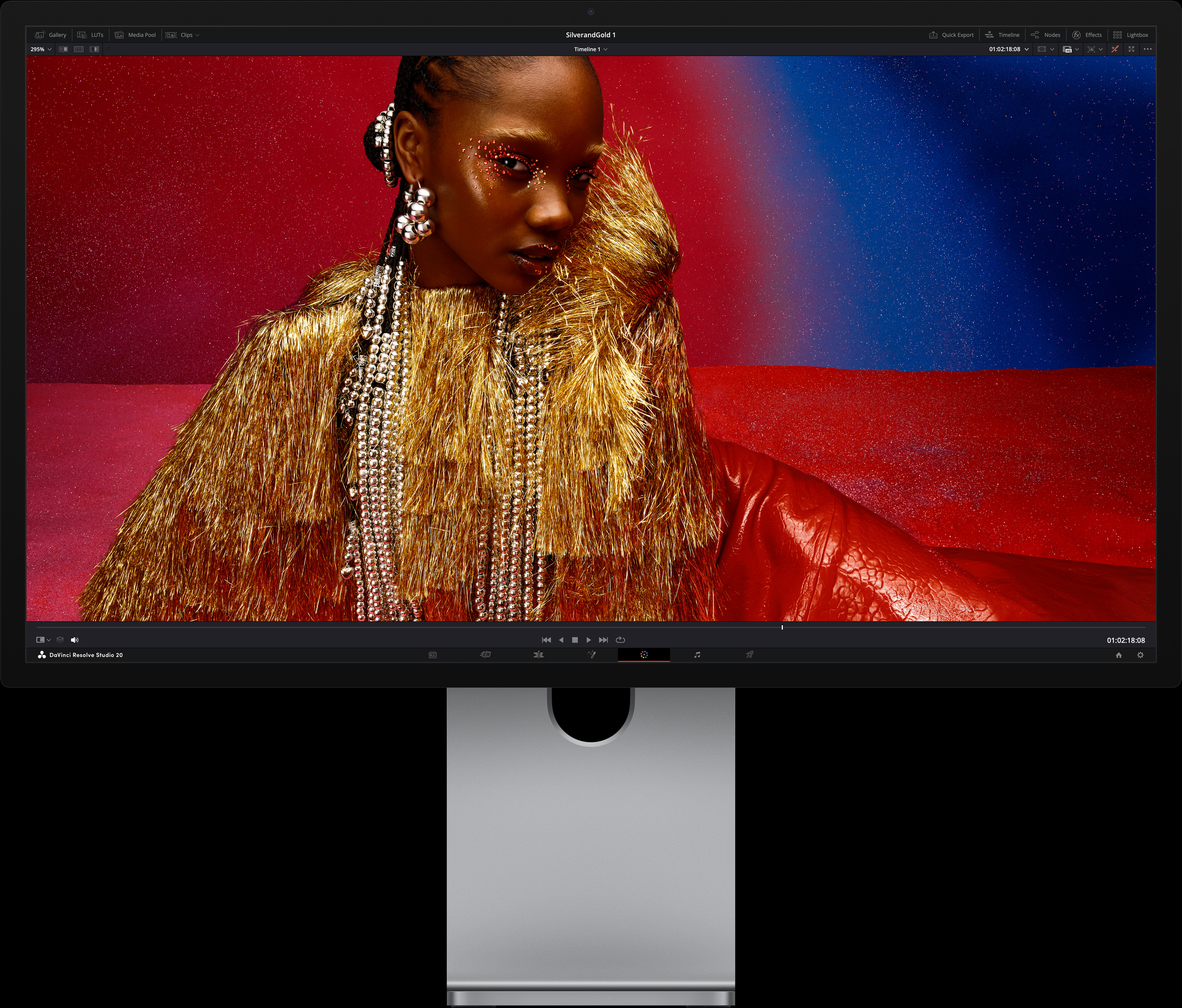 Egy élénkvörös ruhát és túlméretezett fémes kiegészítőket viselő személy egy kék-piros átmenetes háttér előtt ül, bemutatva a Studio Display XDR képminőségét