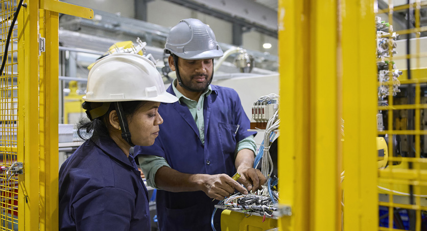 Two employees in work smocks and safety hats stand in front of a assembly machine